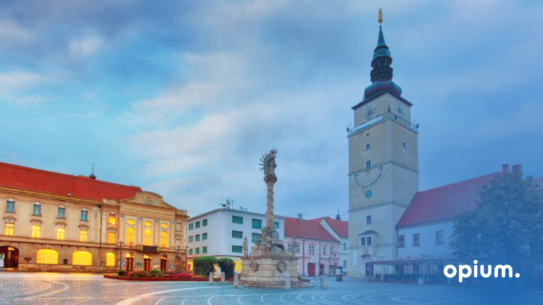 Evropské náměstí za soumraku s historickými budovami, vysokou věží s hodinami a centrálním kamenným památníkem. V pravém dolním rohu je na modrém pozadí s přechodem barev zobrazeno bílé slovo „opium“.