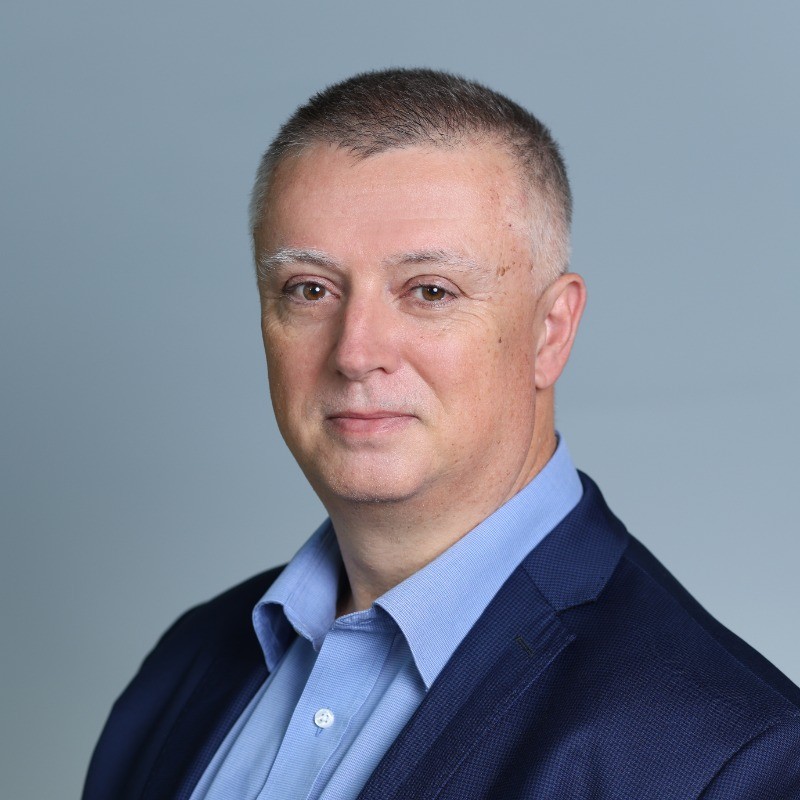 Muž stredného veku s krátkymi šedivými vlasmi, oblečený v modrom saká a svetlomodrej košeli, pózuje pred jednoliatym svetlomodrým pozadím a s neutrálnym výrazom hľadí priamo do kamery.